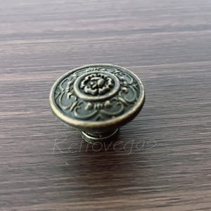 May include: Antique brass-finished cabinet knob with an ornate floral design. The round knob features detailed scrollwork and a raised center, perfect for adding a vintage touch to furniture. The knob is shown on a wooden surface.