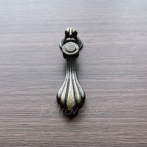 May include: A vintage-style, ornate, black metal cabinet pull with a decorative, fluted design. The pull has a round, circular knob at the top.