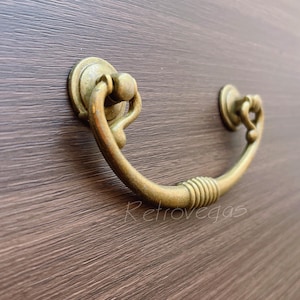 Op de afbeelding: Een gouden metalen ladegreep met een gebogen, sierlijk ontwerp. De greep heeft een gladde, gepolijste afwerking en is geschikt voor gebruik op kasten, ladekasten en andere meubels.