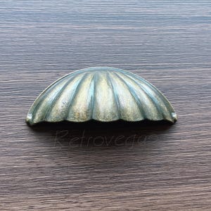May include: A vintage-style, shell-shaped drawer pull with a bronze finish. The hardware has a ribbed design and is set against a dark wood grain background. The word "Retrovega" is visible in the image.