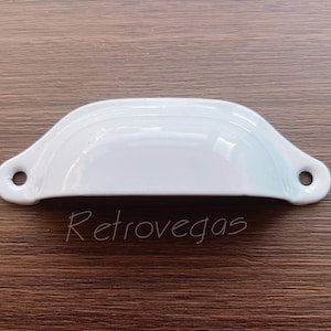 Könnte beinhalten: Weißer Keramik-Schubladengriff mit abgerundeter, geschwungener Form. Der Griff hat eine glatte, glänzende Oberfläche.