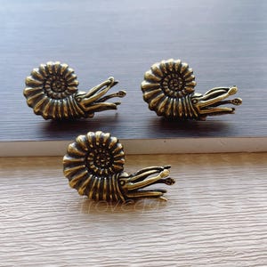 May include: Three bronze-coloured, nautilus-shaped decorative hardware pieces. Each piece features a spiralled shell design with detailed tentacles extending outwards. The hardware is likely for furniture or decorative purposes.