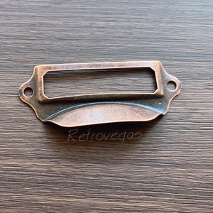 Peut inclure: Une tirette de tiroir en métal de couleur cuivre avec un porte-étiquette rectangulaire. Le porte-étiquette a un dessus légèrement incurvé et des coins arrondis.