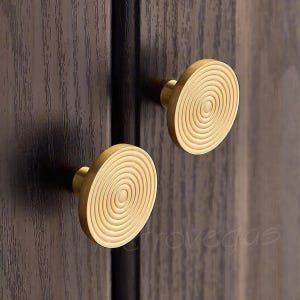 Könnte beinhalten: Zwei goldfarbene runde Schrankknöpfe mit konzentrischem Kreisdesign. Die Knöpfe sind auf einer dunklen Holzoberfläche montiert.