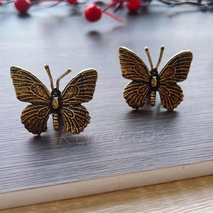 May include: Two antique gold-colored butterfly-shaped decorative objects. The butterflies have detailed wing patterns and are positioned on a gray surface. Red berries and greenery are in the background.