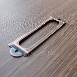 May include: A rectangular, antique-style label holder with a copper finish. The holder has a cutout for a label and two small holes for mounting. The item is set against a dark wood-grain surface.