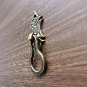 May include: A decorative bronze cabinet pull with a unique, ornate design. The pull features a ring-shaped handle and a detailed, leaf-like shape.