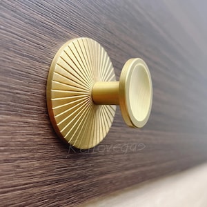 May include: A gold-coloured cabinet knob with a circular face featuring a sunburst pattern. The knob is attached to a dark brown wooden surface. It has a cylindrical stem and a rounded, flat end.