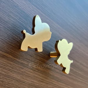 May include: Two gold-colored cabinet knobs in the shape of a dog. The knobs are made of metal and have a smooth finish. The knobs are mounted on a brown wooden surface. The word "Retrovegas" is printed on the surface.
