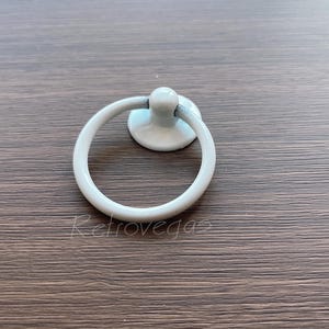 Op de afbeelding: Witte keramische ringgreep voor een kastdeur. De greep is een eenvoudige ringvorm met een ronde basis.