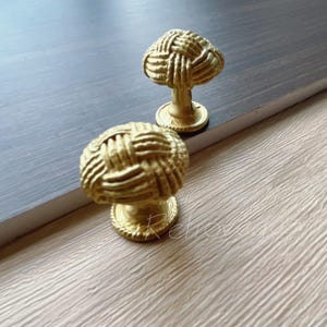 Può includere: Due pomelli per armadi dorati con un design intrecciato. I pomelli sono rotondi e hanno una superficie testurizzata in rilievo.