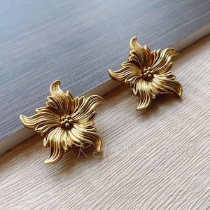 May include: A pair of gold-tone flower-shaped earrings. The earrings are made of metal and have a detailed design.