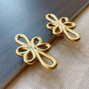 May include: Two gold-toned decorative drawer pulls in the shape of a stylized knot. The pulls are made of metal and have a smooth, polished finish.