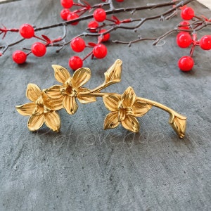 May include: Gold-toned metal flower brooch with two blossoms and a stem. The brooch is on a gray textured surface.