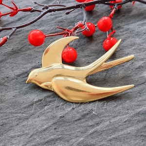 Op de afbeelding: Een gouden metalen zwaluw broche. De broche is simpel van ontwerp met een gladde, glanzende afwerking.