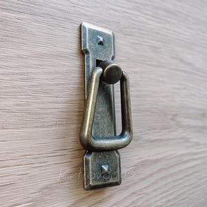 Puede incluir: Un tirador de armario de estilo vintage en latón antiguo con un asa de anillo triangular. El tirador tiene un acabado desgastado y está montado en una superficie de madera.