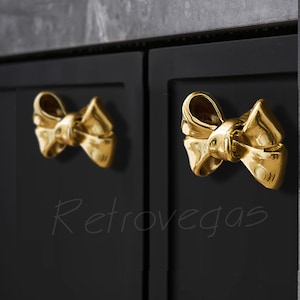 May include: Two gold bow-shaped cabinet knobs on black cabinet doors. The knobs are made of solid brass and have a polished finish.