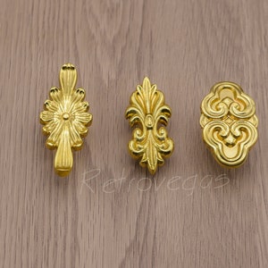 May include: Three gold-toned decorative cabinet knobs with ornate designs. The knobs feature a floral design, a fleur-de-lis design, and a cloud design.