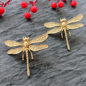 Könnte beinhalten: Ein Paar goldfarbene Libellenohrringe. Die Libellen haben detaillierte Flügel und Körper.