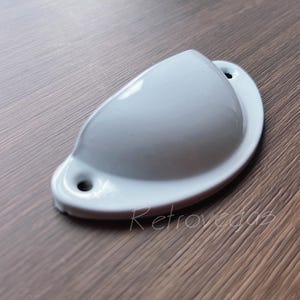 May include: A white, shell-shaped drawer pull with two screw holes. The hardware has a glossy finish and a rounded, ergonomic design. The pull is set against a dark wood grain surface.