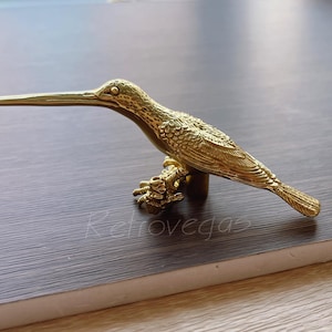 以下が含まれることがあります： 枝にとまった真鍮製のハチドリの彫刻。鳥は長く細い嘴と質感のある羽を持っています。彫刻は装飾品で、おそらく室内装飾用です。
