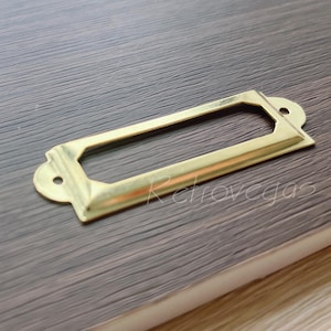 Puede incluir: Un portaetiquetas de metal de color dorado con una abertura rectangular. El portaetiquetas tiene un borde ligeramente redondeado y un acabado liso.
