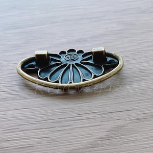 May include: A bronze-coloured drawer pull with an ornate, black floral design. The pull is oval-shaped and has two small, square-shaped mounting plates.