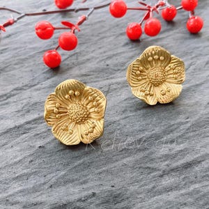 May include: A pair of gold-toned flower-shaped earrings. The earrings are made of metal and have a detailed design.