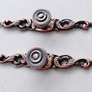 Peut inclure: Deux poignées de tiroir en cuivre ornées d'un motif de volutes décoratives et d'un bouton rond. Les boutons présentent un motif de cercles concentriques.