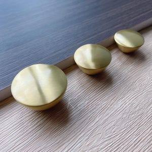 May include: Three brass cabinet knobs with a brushed finish are arranged on a light wood-grain surface next to a grey-blue surface. The knobs are round with a slightly domed shape, suitable for furniture hardware.