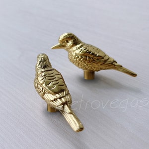 Puede incluir: Dos tiradores de cajón en forma de pájaro de color dorado. Los pájaros están mirando en direcciones opuestas. Los pájaros tienen alas y picos detallados.