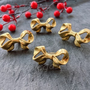 May include: Four gold-coloured bow-shaped napkin rings. The rings are arranged on a grey surface.