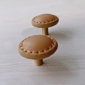May include: Two brown leather cabinet knobs with gold metal bases. The knobs have a stitched edge.
