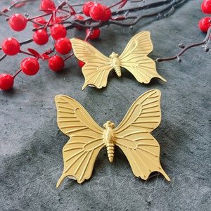 Peut inclure: Deux ornements en métal en forme de papillon dorés sur une surface texturée grise. Les papillons ont des ailes détaillées et une finition texturée.