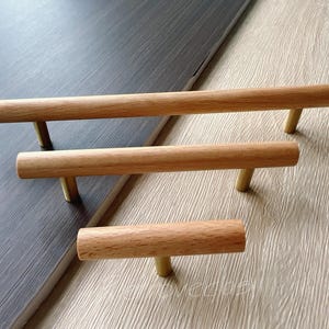 Puede incluir: Tres tiradores de madera para armarios con vástagos de color latón. Los tiradores cilíndricos son de madera clara. Están dispuestos sobre una superficie de vetas de madera gris oscuro y beige claro.