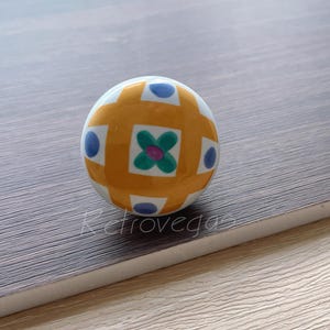 May include: A decorative ceramic ball with a geometric pattern. The ball features a white base with orange, blue, and green accents. The design includes squares, dots, and a floral motif. The ball is resting on a wooden surface.