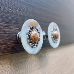 May include: Two oval cabinet knobs with a mother-of-pearl finish and ornate silver filigree. Each knob has a brown, reflective stone centre. The knobs are mounted on a dark wood surface.