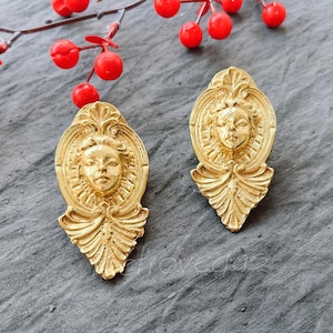 May include: A pair of gold-tone earrings with a detailed design featuring a woman's face. The earrings are shaped like a teardrop and have a textured surface.