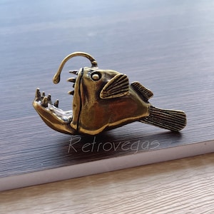 Pode incluir: Uma figurinha de peixe-pescador de cor bronze com uma grande boca aberta e uma isca na parte superior da cabeça. A figurinha é detalhada com escamas e barbatanas texturizadas.