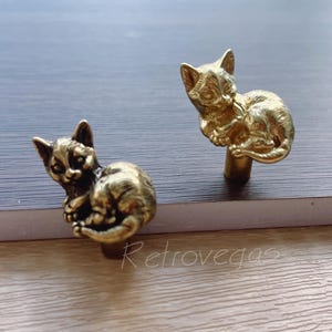 Puede incluir: Dos figuritas de gato de metal dorado, cada una con un gatito enroscado en su espalda. Los gatos están detallados con rasgos realistas y están diseñados para ser utilizados como tiradores de cajones o pomos de armario.