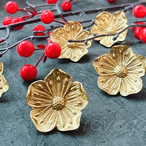 May include: Five gold metal flower-shaped decorative pieces with a textured surface. The flowers are arranged on a grey fabric background.