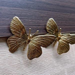 Może przedstawiać: Dwa złote uchwyty do szafek w kształcie motyla. Uchwyty motylkowe są wykonane z metalu i mają szczegółowe wzory skrzydeł. Uchwyty są zamontowane na drewnianej powierzchni z jasnym usłojeniem.