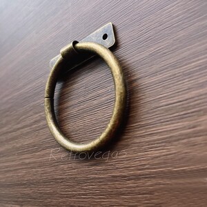 Può includere: Un anello di metallo dorato con una piastra rettangolare di metallo fissata sul retro. L'anello è montato su una superficie di legno marrone.