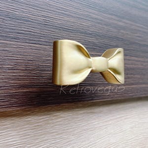 May include: A gold-colored bow-shaped cabinet pull. The hardware is mounted on a dark brown wooden surface. The word "Retrovegas" is printed in a cursive font on the lower left side of the image.