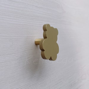 以下が含まれることがあります： 金色のテディベア型のキャビネットノブ。白い木製の表面に取り付けられています。テディベアのデザインは家具に遊び心を加えます。ノブは金属製です。