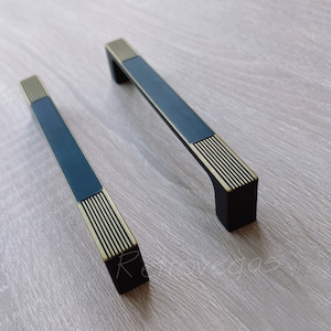 Może przedstawiać: Dwa czarne uchwyty do szafek z złotymi akcentami. Uchwyty mają prostokątny kształt z płaską powierzchnią i żłobionym wzorem.