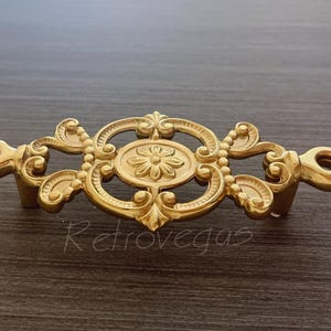 May include: Gold-toned metal drawer pull with an ornate design featuring a central flower motif and scrollwork.