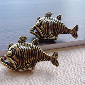 May include: Two gold-toned metal fish-shaped drawer pulls. The fish have a detailed, textured surface and are facing the same direction.