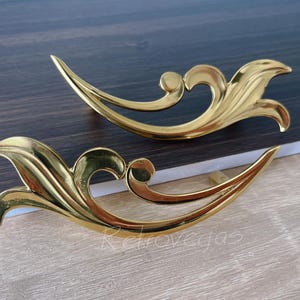 May include: Two gold-colored, ornate cabinet pulls with a flowing, leaf-like design. The pulls have a curved shape with a central swirl and pointed ends. The hardware is set against a dark wood surface.