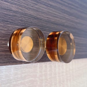May include: Two cabinet knobs with a clear, rounded design. The knobs feature a circular, gold-colored center and are encased in a clear material with alternating bands of brown and amber. The knobs are mounted on a dark wood surface.